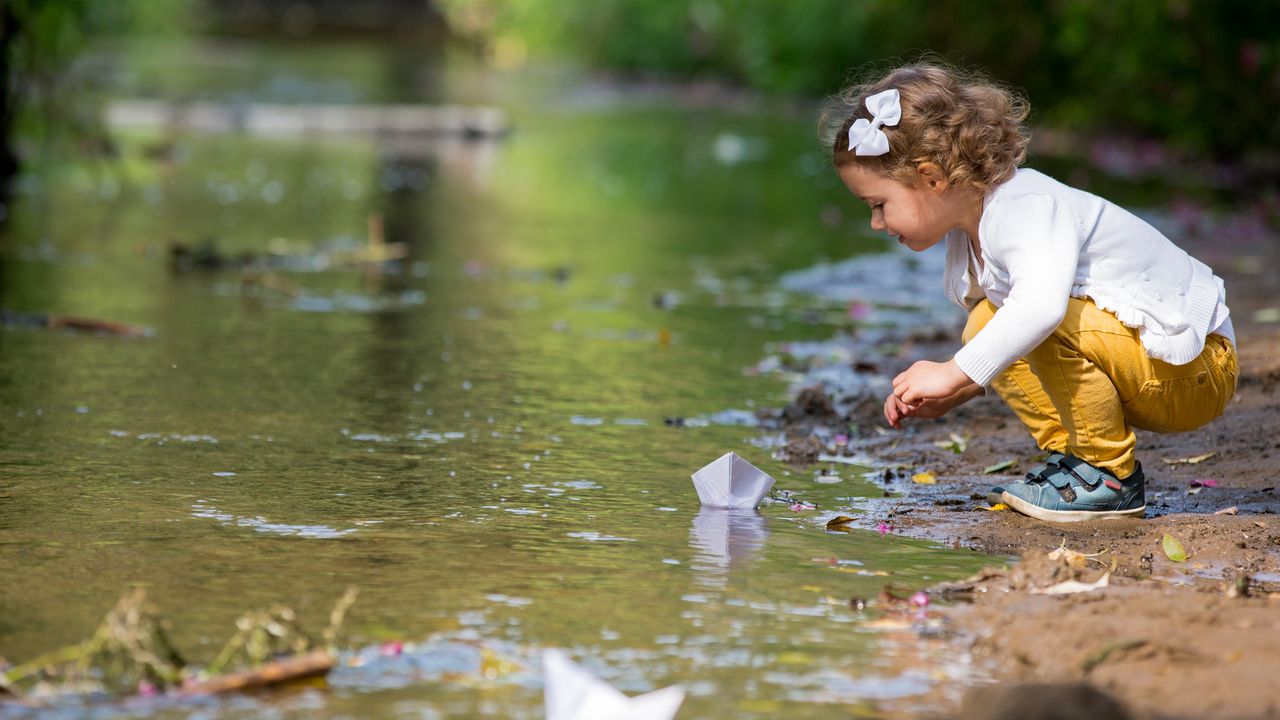 Ein kleines Kind, welches mit einem Papierschiff am Flussufer spielt. 